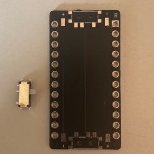 May include: A black circuit board with rows of round holes and a small toggle switch. The circuit board has the letters "D7" and "B10" printed on it.