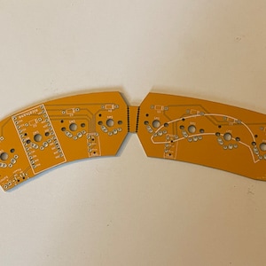 May include: A two-piece, orange-colored circuit board with white text and markings. The board is curved and has a series of small holes.