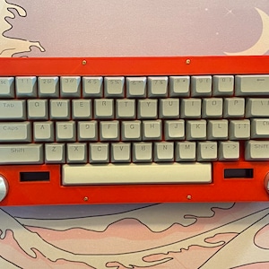 Könnte beinhalten: Eine rote mechanische Tastatur mit grauen Tastenkappen und einer weißen Leertaste. Die Tastatur hat eine rechteckige Form mit abgerundeten Ecken und zwei silbernen Knöpfen an den Seiten. Die Tasten sind mit weißen Buchstaben und Symbolen beschriftet.