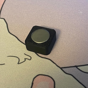 May include: A small, square, black object with a round, silver button in the center.