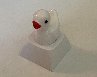 May include: A white duck-shaped artisan keycap with a red beak and black eyes. The keycap is mounted on a white, rectangular base, creating a whimsical design. This custom keycap is a unique accessory for mechanical keyboards.