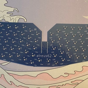 May include: A blue printed circuit board with a split design and the text "atreus62" printed on it. The board has many small holes and silver-colored components.