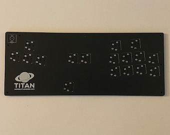 May include: Black keyboard plate with a white logo that says "TITAN WEIRD LITTLE KEEDS.COM" and a white planet symbol. The plate has 18 holes for mechanical keyboard switches.