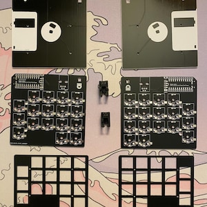 Könnte beinhalten: Ein Satz schwarzer Kunststoffkomponenten zum Bau einer individuellen Tastatur. Das Set enthält zwei schwarze Kunststoffplatten, zwei schwarze Kunststoffrahmen, zwei schwarze Kunststoffkappen und zwei schwarze Kunststoffschalter. Die Komponenten sind auf einer rosa-weißen gemusterten Oberfläche angeordnet.