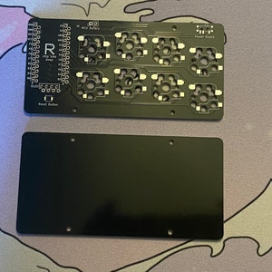 May include: A black circuit board with a row of buttons and a reset button labeled "Reset Button". The board has text that reads "MCU Battery" and "Power Switch". There is a second black board with no features.