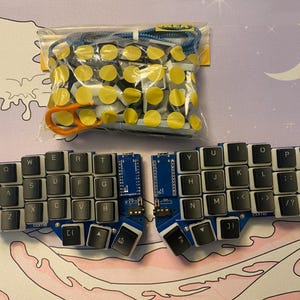 Puede incluir: Un teclado dividido con teclas negras y letras blancas. El teclado tiene una placa de circuito azul y negra. El teclado está sobre un fondo rosa y blanco.