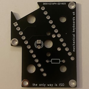May include: Black circuit board with silver components and the text "mechanical keyboards uk" and "the only way is ISO". The board has a unique shape with multiple holes and connection points.