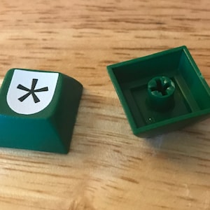 May include: A green keyboard key with a white sticker featuring a black asterisk symbol. The key is shown from the top and bottom.