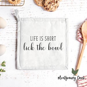 May include: White cotton pot holder with the text "Life is short, lick the bowl" in black script. The pot holder is on a white wooden surface with a red and white striped tea towel, a wooden spoon, and a few cookies on a wire rack.