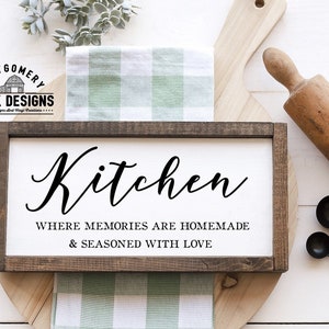 May include: A wooden sign with the text "Kitchen, Where memories are homemade & seasoned with love". The sign is on a white surface with a green and white checkered cloth and a wooden rolling pin.