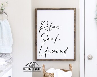 Puede incluir: Un cartel blanco enmarcado con caligrafía negra que dice "Relax Soak Unwind". El cartel está colgado en una pared blanca en un baño.