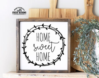 以下が含まれることがあります： 茶色の枠が付いた白い木製の看板で、黒いリースの中に「Home sweet home」という文字が筆記体で書かれています