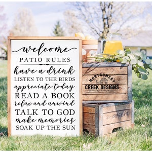 May include: A white wooden sign with black text that reads "welcome patio rules have a drink listen to the birds appreciate today read a book relax and unwind talk to god make memories soak up the sun". The sign is leaning against a wooden crate with the text "Montgomery Creek Designs" on it.