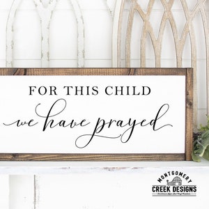 Può includere: Un cartello di legno con il testo "For this child we have prayed" in corsivo nero. Il cartello è incorniciato da una cornice in legno marrone scuro. Il cartello è appoggiato su uno scaffale bianco con una parete bianca dietro.