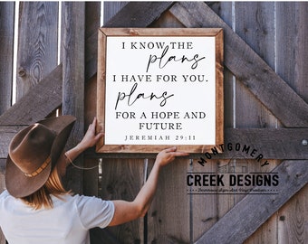 May include: A white wooden sign with a black calligraphy quote, "I know the plans I have for you. Plans for a hope and future. Jeremiah 29:11". The sign is being hung on a wooden fence.