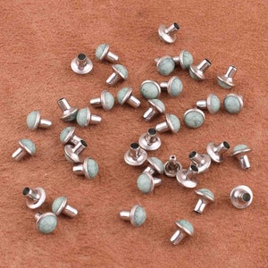 May include: A collection of silver-toned metal studs with green and white marbled tops. The studs are scattered on a brown surface.