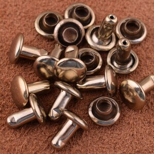 May include: A close-up of a pile of gold-toned metal rivets. The rivets are round with a flat head and a pointed end. They are scattered on a brown leather surface.