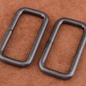May include: Two dark gray metal rectangular rings. The rings have rounded corners and a matte finish. The rings are placed on a brown surface. These rings could be used for bag hardware or other craft projects.