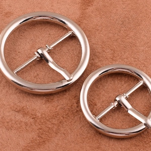 May include: Two silver metal belt buckles, one larger than the other, on a brown suede surface.