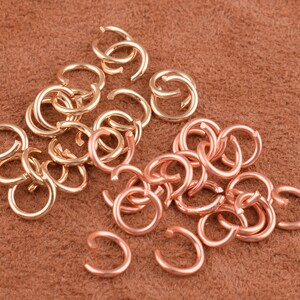 May include: A collection of gold and copper jump rings. The jump rings are small and round, and they are arranged in a pile on a brown surface.