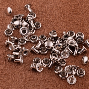 May include: A large pile of silver metal rivets, some with a raised dome, on a brown leather surface.