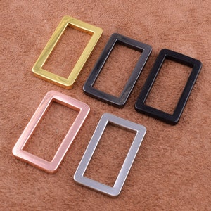 May include: Five metal belt buckle loops in different finishes: gold, silver, black, and rose gold. The loops are rectangular with rounded corners.