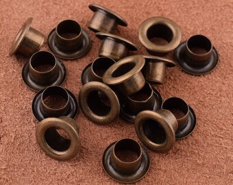May include: A collection of antique bronze-colored grommets, each with a circular eyelet and a wide, flat flange. The grommets are scattered across a textured, brown surface, showcasing their aged appearance and metallic sheen. Ideal for crafting and leatherwork.