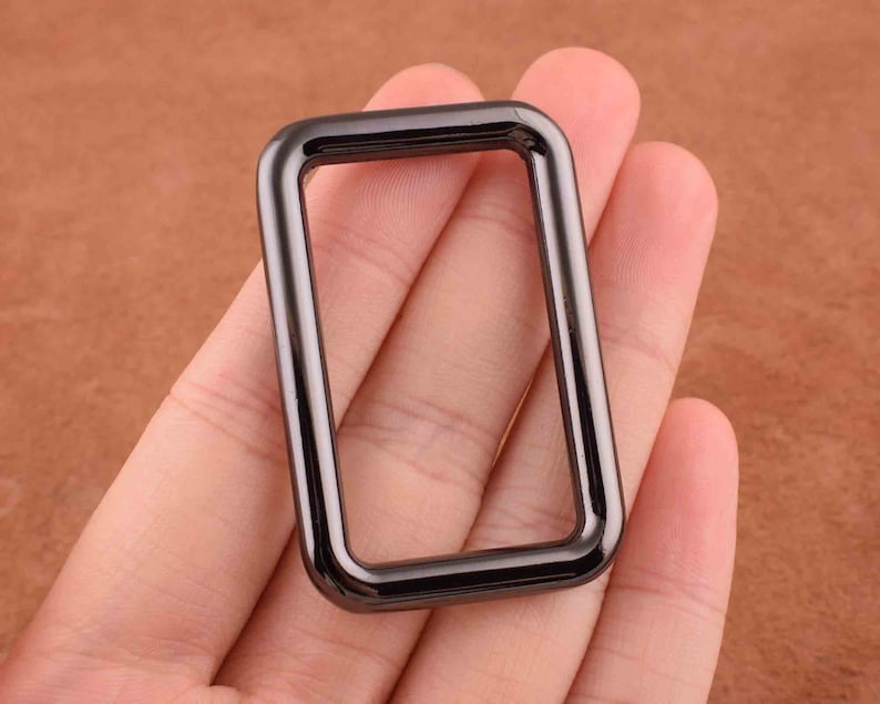 Peut inclure: Un anneau en D en m&eacute;tal noir avec une finition lisse. L'anneau est de forme rectangulaire et a des coins arrondis.