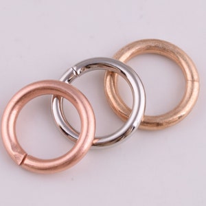 Può includere: Tre anelli metallici di diversi colori: oro, argento e rame. Gli anelli sono tutti di dimensioni simili e sono appoggiati su una superficie bianca.