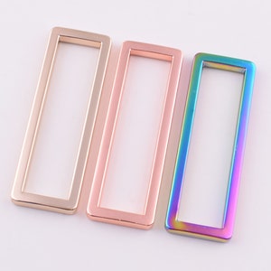 Puede incluir: Tres anillos rectangulares de metal en oro, oro rosa y colores arcoíris iridiscentes. Los anillos están vacíos en el centro y tienen un acabado liso y pulido. Estos anillos decorativos probablemente se utilizan para manualidades o joyería.