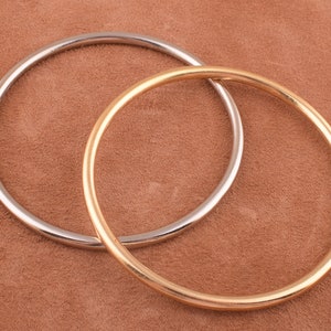 Pode incluir: Dois anéis de metal, um dourado e outro prateado, estão sobre uma superfície marrom. Os anéis são simples e circulares.