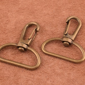 May include: Two antique gold-toned metal swivel clasps with a D-ring. The clasps are open and ready to be attached to a bag or other item.