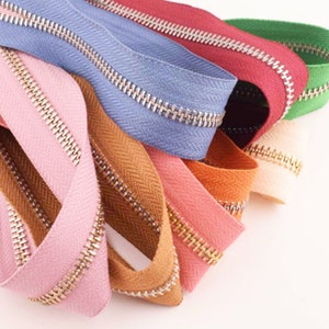 Może przedstawiać: Zestaw kolorowych taśm suwakowych ze złotymi metalowymi zębami. Taśmy są ułożone wachlarzowo, prezentując różne kolory i faktury.