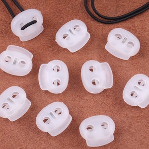 May include: Ten white plastic cord stoppers with two holes each. The stoppers have a smooth, rounded surface and are designed to secure cords or straps.