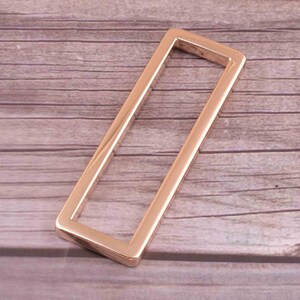 May include: A rose gold metal rectangular frame with rounded corners. The frame is empty and has a smooth, polished finish.