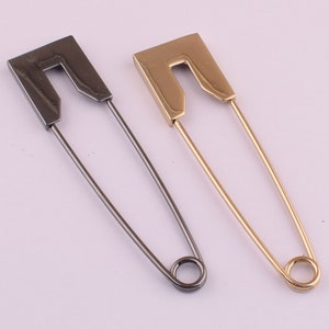 May include: Two safety pins, one gold and one black, are shown against a white background. The pins are both closed and have a simple, rectangular design.