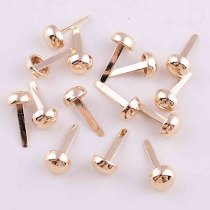 May include: A collection of gold-toned metal brads with round heads and straight pins. The brads are scattered on a white surface.