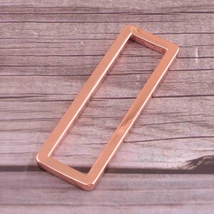 May include: A rose gold metal rectangle buckle with rounded corners. The buckle is designed to be used for straps or belts.