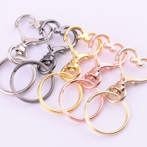 Könnte beinhalten: Vier herzförmige Schlüsselanhänger in verschiedenen Metalloberflächen: Silber, Schwarz, Gold und Roségold. Jeder Schlüsselanhänger hat einen runden Ring und einen drehbaren Karabinerhaken.