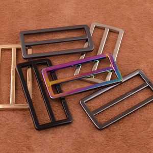 May include: Five metal belt buckle sliders in different finishes. The buckles are rectangular with two horizontal bars. The finishes include gold, silver, black, and iridescent.