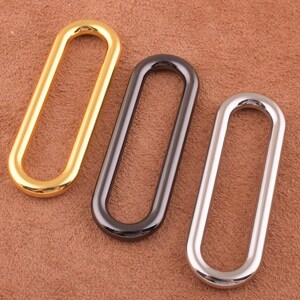 May include: Three oval-shaped metal rings in gold, black, and silver. The rings are smooth and have a polished finish.