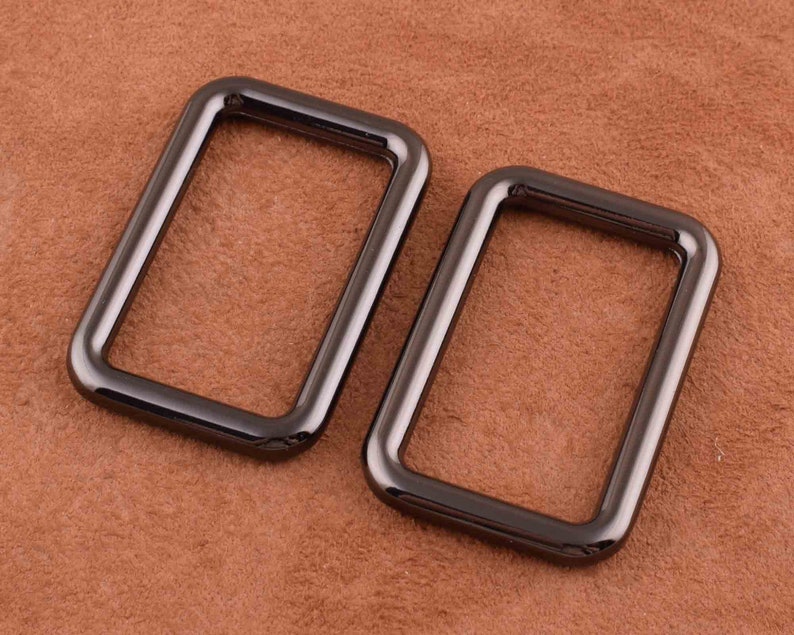 Peut inclure: Deux anneaux en D en m&eacute;tal noir, chacun de forme rectangulaire et &agrave; la finition lisse et polie. Les anneaux sont pos&eacute;s sur une surface en cuir brun.