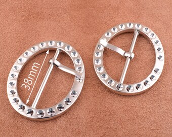 May include: Two silver metal belt buckles with a circle of small raised dots around the edge. The buckle on the left has a measurement of 38mm marked on it.