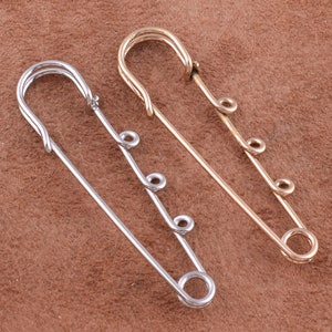 May include: Two safety pins, one silver and one gold, with multiple loops on the pin. The pins are on a brown textured surface.
