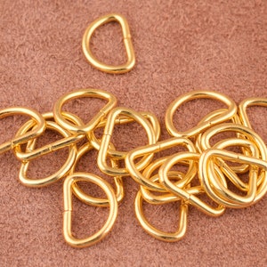 May include: A close-up of a pile of gold-colored metal D-rings. The rings are all the same size and are arranged in a random pattern.