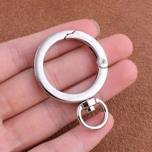 May include: A silver-colored metal clasp with a large circular ring and a smaller ring attached. The clasp is open, revealing the hinge mechanism. The background is a brown, textured surface.