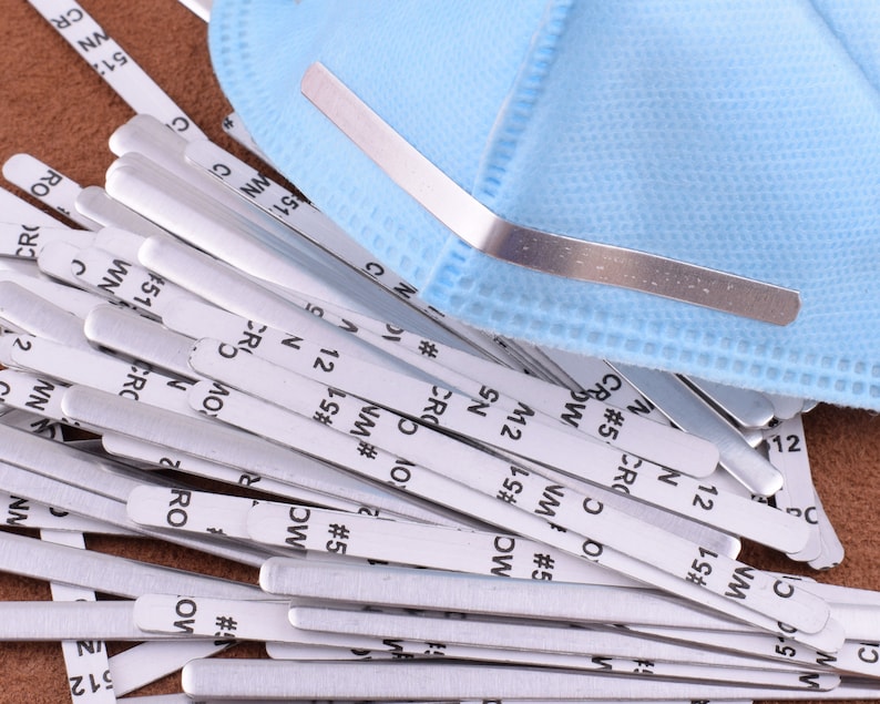 May include: A close-up of a pile of silver nose wire inserts for face masks. The wires are labeled with numbers and letters, such as "#5", "NM", and "CR".