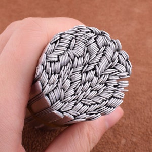 May include: A close-up of a stack of silver metal rings, woven together in a basket weave pattern.