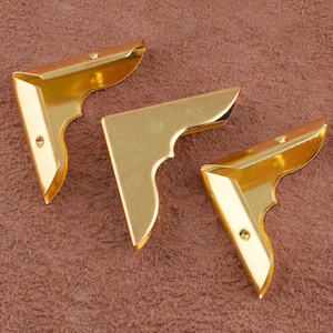 May include: Three gold metal corner protectors with a decorative design. The corner protectors are on a brown surface.