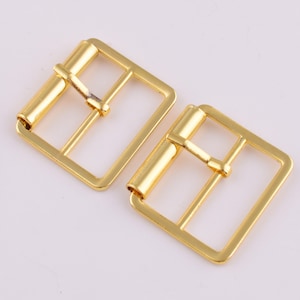 May include: Two gold-toned metal belt buckles. Each buckle has a square shape with a rectangular opening in the center. The buckles are designed to be used with a belt.